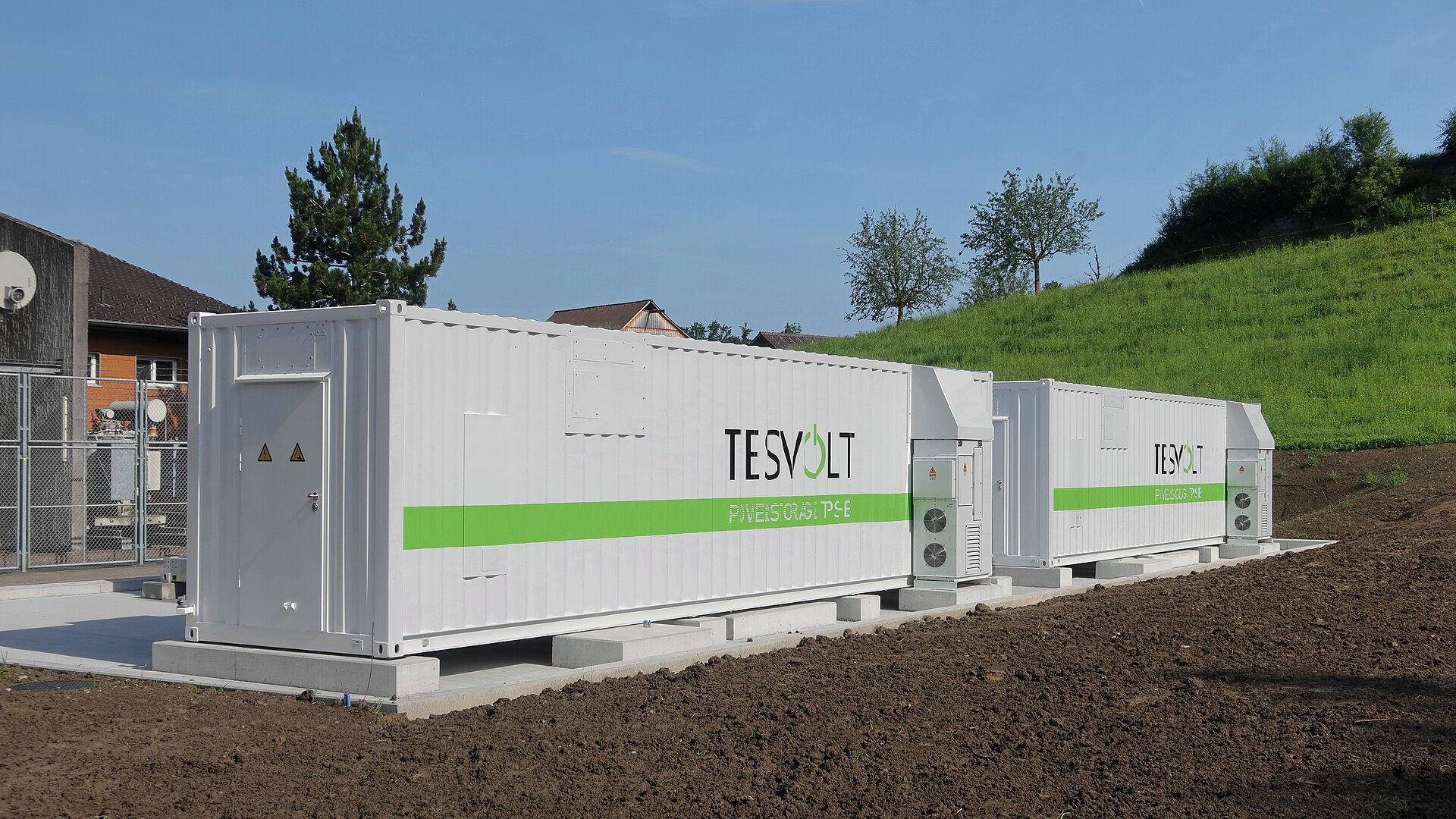 Two white containerised battery energy storage units with cooling units on a concrete foundation beside a grid substation